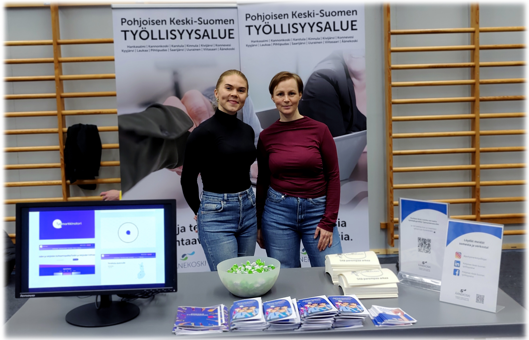 Kaksi naista seisoo pöydän takana. Pöydällä on esittelyaineistoa ja karkkeja. Taustalla on kaksi isoa julistetta, joissa lukee Pohjoisen Keski-Suomen työllisyysalue.