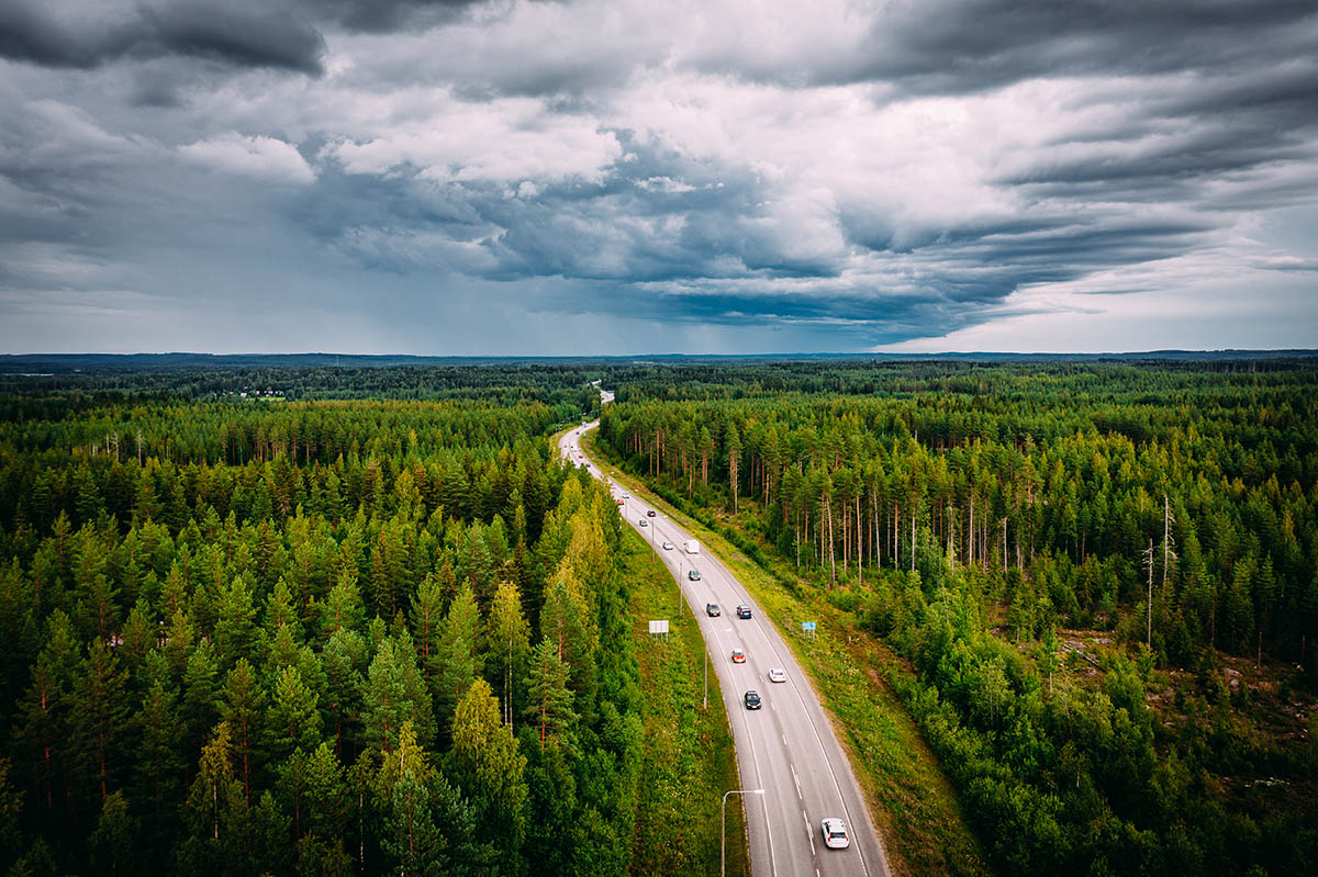 Kuvassa on pilvistä taivasta ja metsän keskellä tie, jolla ajaa autoja.