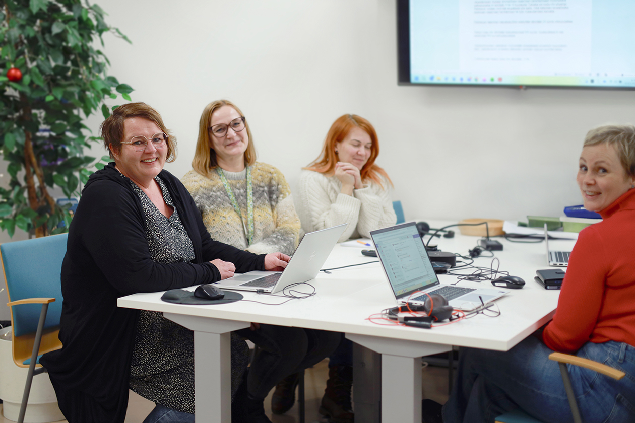 Pöydän äärellä istuu neljä naista. Kolme heistä katsoo kameraan ja yksi hymyilee silmät kiinni. Pöydällä on kannettavia tietokoneita ja muita pieniä toimistotarvikkeita.